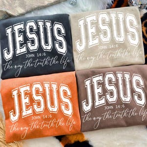 May include: Four sweatshirts in different colours: black, beige, orange, and brown. Each has the word "JESUS" in large, white, collegiate-style lettering. Below the word "JESUS" is the text "JOHN 14:6" and "the way the truth the life".