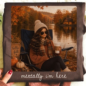 May include: A dark brown t-shirt featuring a printed graphic of a person in a knit beanie, sunglasses, and scarf sitting in a folding chair by a lake with autumn trees. The text "hello fall" is on a sign, and "mentally i'm here" is printed below.