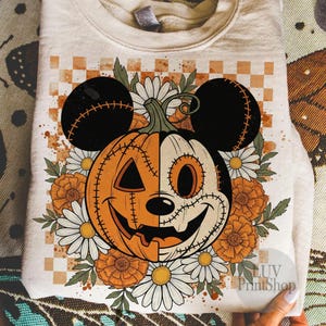 Puede incluir: Una sudadera color crema con un diseño temático de Halloween. El gráfico representa una calabaza con orejas de Mickey Mouse, rodeada de flores. El diseño es naranja, negro y blanco. El texto "LUV PrintShop" es visible.