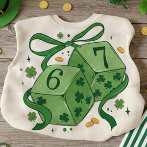 Puede incluir: Una sudadera blanca con una cinta verde y dos dados verdes, uno con el número 6 y otro con el número 7. Los dados están decorados con tréboles. Monedas de oro y un sombrero verde están dispersos.