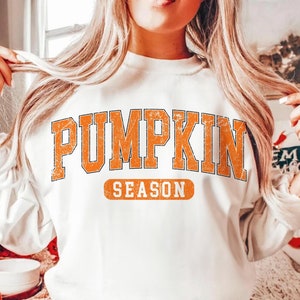 May include: White sweatshirt with the words "PUMPKIN SEASON" in orange, distressed lettering. The sweatshirt has a crew neck and long sleeves. The overall theme is autumn and fall.
