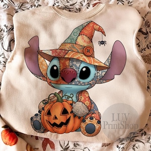 Könnte beinhalten: Ein cremefarbenes Sweatshirt mit einer Cartoonfigur, die einen Patchwork-Hexenhut trägt und einen orangefarbenen Kürbis hält. Die Figur ist blau mit großen schwarzen Augen und einer rosa Nase. Eine kleine Spinne hängt am Hut. Der Text "LUV PrintShop" ist sichtbar.