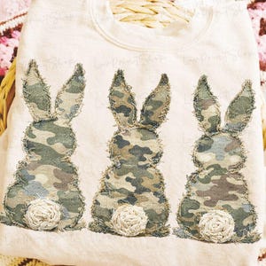 Op de afbeelding: Een crèmekleurige sweatshirt met drie geappliqueerde konijnen. De konijnen hebben camouflage patronen op hun lichaam en oren, met witte, pluizige staartaccenten. De sweatshirt is waarschijnlijk een seizoensgebonden of feestelijk item.