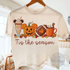 May include: A cream-coloured t-shirt with a seasonal graphic. The design features a pumpkin with Mickey Mouse ears, a football with a Mickey Mouse logo, a milkshake, a mug of hot chocolate, and the text "Tis the season."