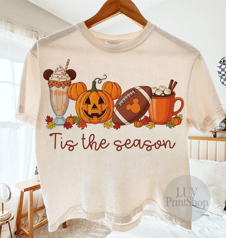 May include: A cream-coloured t-shirt with a seasonal graphic. The design features a pumpkin with Mickey Mouse ears, a football with a Mickey Mouse logo, a milkshake, a mug of hot chocolate, and the text "Tis the season."
