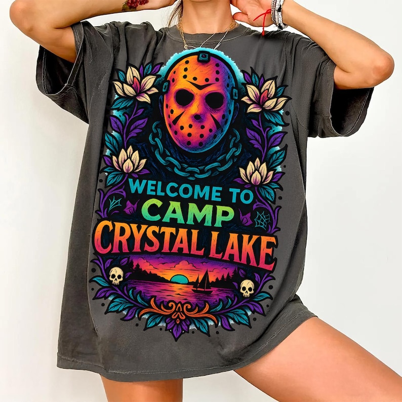 May include: A charcoal gray t-shirt featuring a colorful graphic of a hockey mask, chains, and floral elements. The text reads WELCOME TO CAMP CRYSTAL LAKE above a sunset lake scene with a sailboat and two small skulls at the bottom of the design.