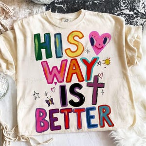 May include: Cream-colored t-shirt with the phrase "HIS WAY IS BETTER" in colorful, hand-painted lettering. A pink heart with a smiling face, a cross, a butterfly, and other small illustrations are also featured.