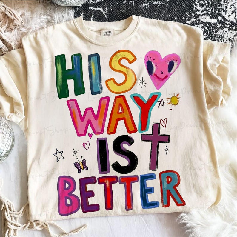 May include: Cream-colored t-shirt with the phrase "HIS WAY IS BETTER" in colorful, hand-painted lettering. A pink heart with a smiling face, a cross, a butterfly, and other small illustrations are also featured.