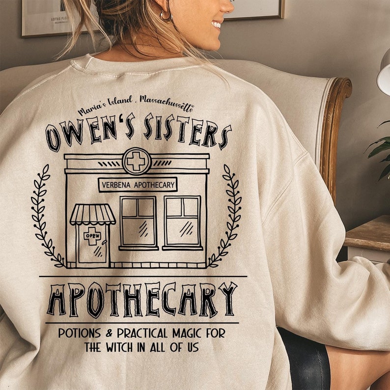 以下が含まれることがあります： ベージュのスウェットシャツ。黒いグラフィックデザインで、「Owen's Sisters Apothecary」と「Maria's Island, Massachusetts」の文字が入っています。薬局の建物のイラストと「Potions & Practical Magic for the Witch in All of Us」というフレーズも含まれています。