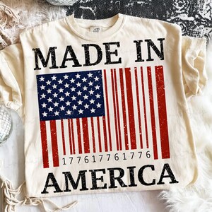 Peut inclure: T-shirt cr&egrave;me avec un motif vieilli du drapeau am&eacute;ricain en forme de code-barres. Les mots "MADE IN AMERICA" sont imprim&eacute;s en noir, avec le code-barres affichant les chiffres "177617761776".