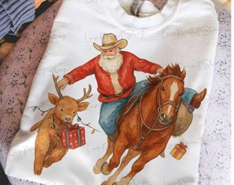 Pode incluir: Camisola branca com uma ilustração em aquarela do Pai Natal montado num cavalo castanho, com uma rena e presentes. O Pai Natal usa um chapéu de cowboy e um casaco vermelho. Roupas com tema natalino.
