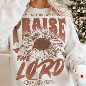 May include: Cream-colored sweatshirt with a large brown sunflower graphic and the words "MY LAST BREATH I WILL RAISE THE LORD" in brown. The text "PSALM 146:2" is printed at the bottom. A cozy and inspirational design.