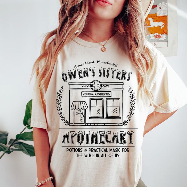 以下が含まれることがあります： クリーム色のTシャツに黒いグラフィックデザイン。デザインには、「OWEN'S SISTERS APOTHECARY」の文字と、店のイラスト、そして「POTIONS & PRACTICAL MAGIC FOR THE WITCH IN ALL OF US」というフレーズが含まれています。