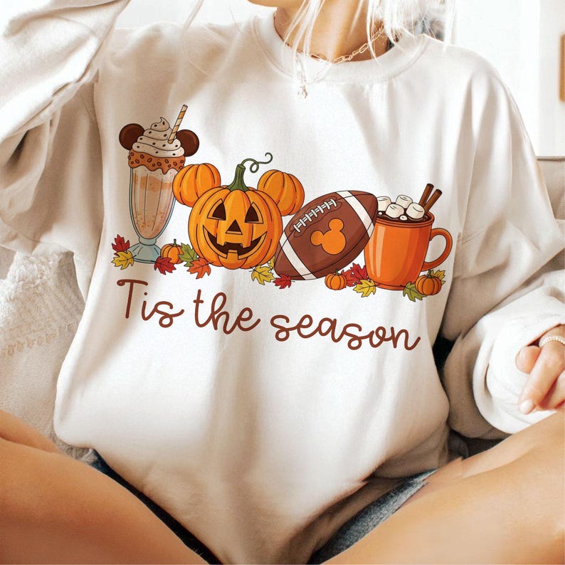 May include: Cream-coloured sweatshirt featuring a seasonal design. The graphic includes a pumpkin with Mickey Mouse ears, a football with a Mickey Mouse logo, a milkshake, and a mug of hot chocolate. The text "Tis the season" is written in a script font.