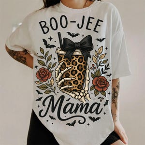Op de afbeelding: Wit T-shirt met een Halloween-thema. Het ontwerp toont de tekst "BOO-JEE Mama" met een skelet hand die een beker met luipaardprint en een zwarte strik vasthoudt. Inclusief vleermuizen, rozen en bladeren.