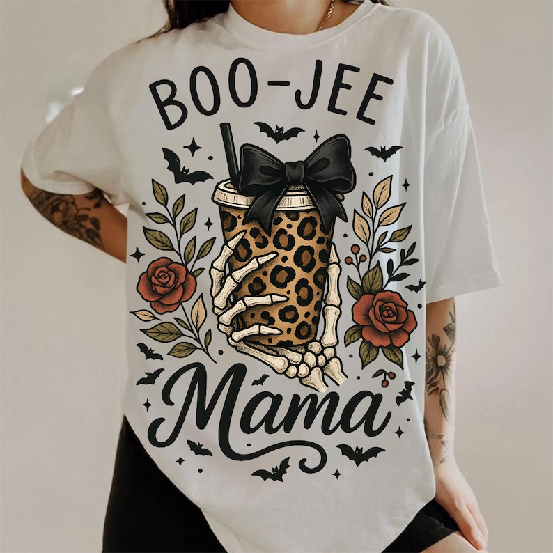 Op de afbeelding: Wit T-shirt met een Halloween-thema. Het ontwerp toont de tekst "BOO-JEE Mama" met een skelet hand die een beker met luipaardprint en een zwarte strik vasthoudt. Inclusief vleermuizen, rozen en bladeren.