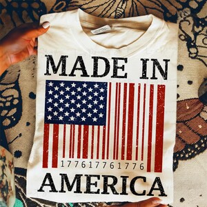 Peut inclure: T-shirt cr&egrave;me avec les mots "MADE IN AMERICA" en lettres noires. Le motif pr&eacute;sente un drapeau am&eacute;ricain stylis&eacute; avec un motif en forme de code-barres. Les &eacute;toiles et les rayures du drapeau sont visibles.
