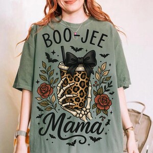 Op de afbeelding: Saliegroen T-shirt met een Halloween-thema. Het ontwerp toont een beker met luipaardprint en een zwarte strik, vastgehouden door een skelet hand, omringd door rozen, bladeren en vleermuizen. De tekst luidt "BOO-JEE Mama".