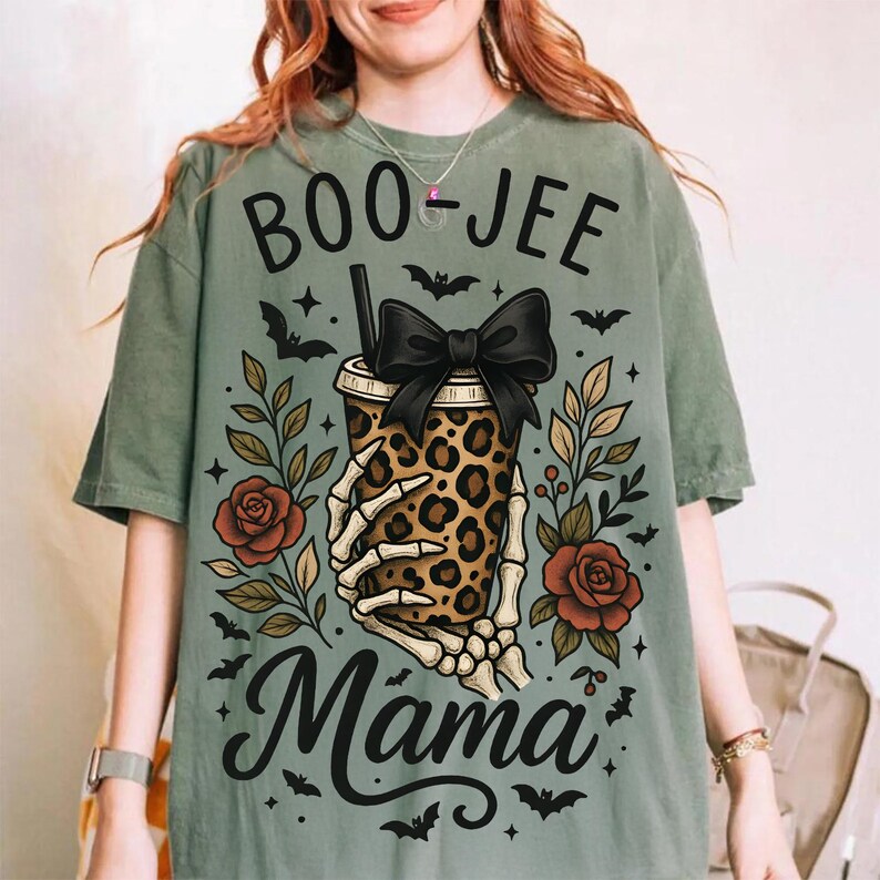 Op de afbeelding: Saliegroen T-shirt met een Halloween-thema. Het ontwerp toont een beker met luipaardprint en een zwarte strik, vastgehouden door een skelet hand, omringd door rozen, bladeren en vleermuizen. De tekst luidt "BOO-JEE Mama".