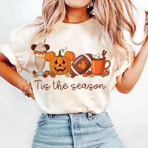 May include: A cream-coloured t-shirt featuring a seasonal graphic. The design includes a milkshake, pumpkins, a football, and a mug of hot chocolate. The text "Tis the season" is written in a script font.