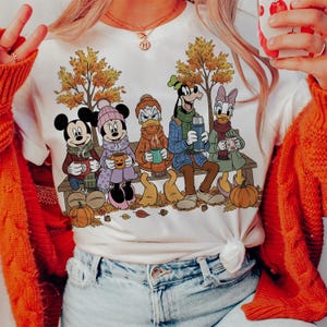May include: White t-shirt featuring Mickey Mouse, Minnie Mouse, Donald Duck, Goofy, and Daisy Duck sitting on a bench in autumn attire. They are holding mugs and a thermos, with pumpkins and fall leaves around them.