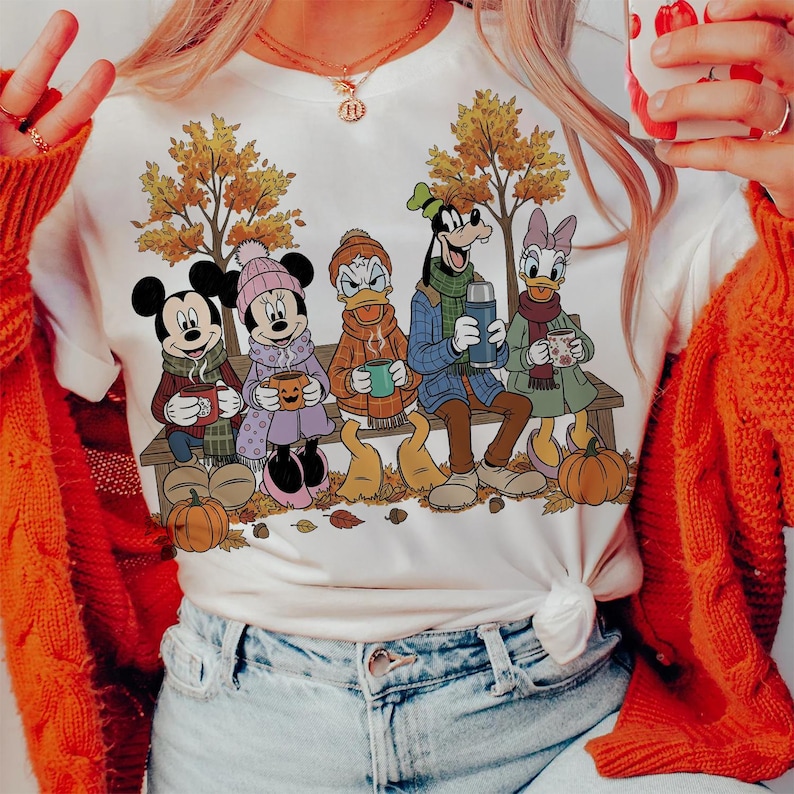 May include: White t-shirt featuring Mickey Mouse, Minnie Mouse, Donald Duck, Goofy, and Daisy Duck sitting on a bench in autumn attire. They are holding mugs and a thermos, with pumpkins and fall leaves around them.