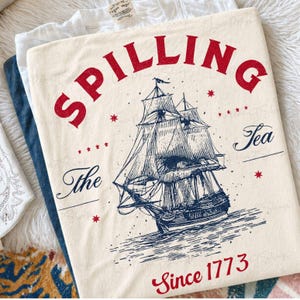 May include: Cream-colored t-shirt with a red and blue graphic of a sailing ship and the words "Spilling Tea Since 1773". The design includes stars and the words "The Tea". The shirt is folded.