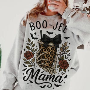Op de afbeelding: Een lichtgrijze sweatshirt met een Halloween-thema. Het ontwerp toont de woorden "BOO-JEE Mama" met een beker met luipaardprint die wordt vastgehouden door een skelet hand, een zwarte strik, vleermuizen en rozen.