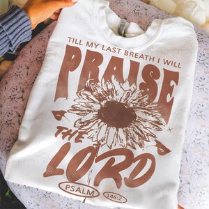 May include: White sweatshirt with a brown sunflower graphic and text that reads "Till my last breath I will praise the Lord" with "Psalm 146:2" below. The sweatshirt is folded on a floral patterned surface.