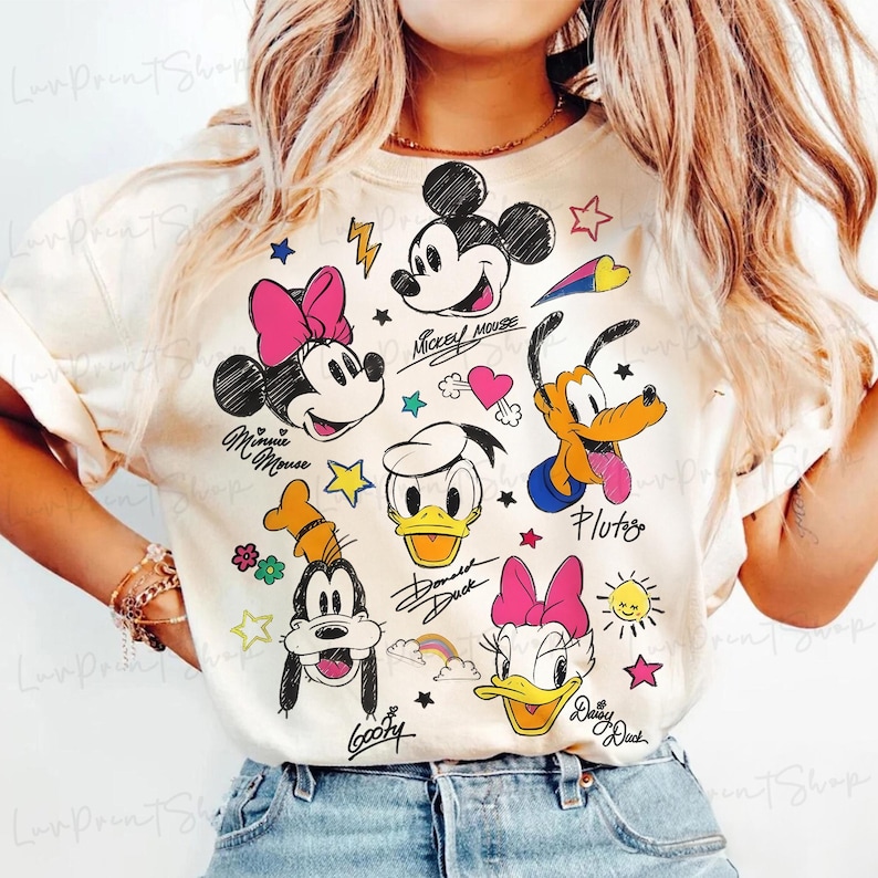 Puede incluir: Camiseta color crema con ilustraciones de dibujos animados de Mickey Mouse, Minnie Mouse, Donald Duck, Daisy Duck, Goofy y Pluto. El dise&ntilde;o incluye estrellas, corazones y los nombres de los personajes. Una camiseta gr&aacute;fica divertida y colorida.