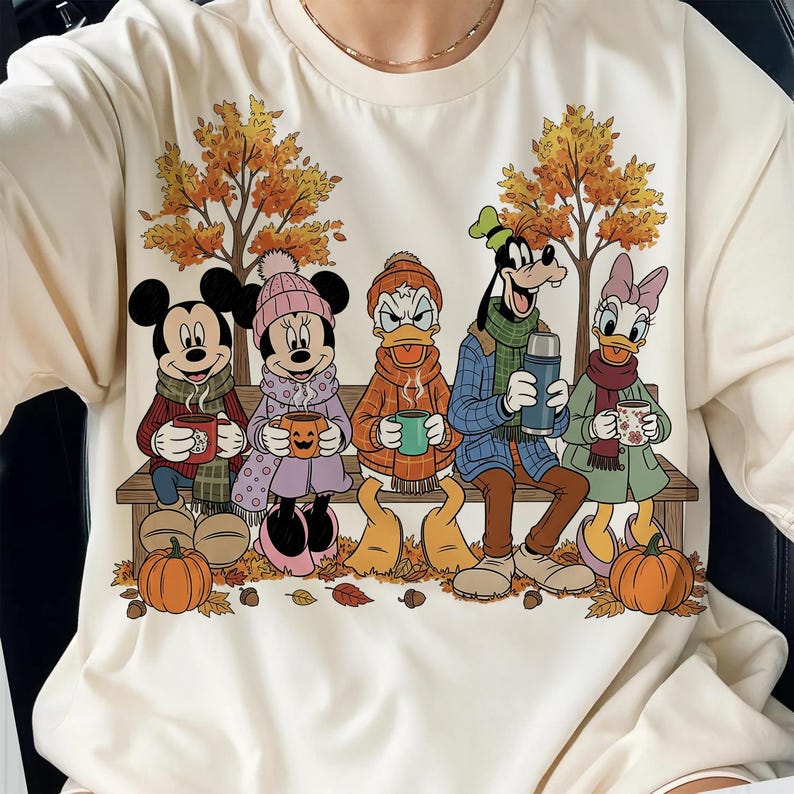 May include: Cream-colored long-sleeved shirt featuring a fall scene with cartoon characters. Mickey and Minnie Mouse, Donald Duck, Goofy, and Daisy Duck are seated on a bench, holding mugs and a thermos. Pumpkins and autumn leaves are scattered around.