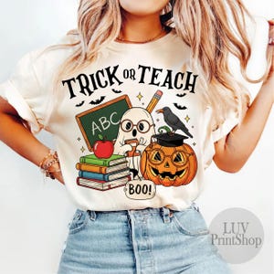 May include: Cream-colored t-shirt with a Halloween-themed graphic. The design features a ghost, a jack-o'-lantern wearing glasses, a chalkboard with "ABC", books, and the text "TRICK OR TEACH". A crow sits atop the pumpkin.