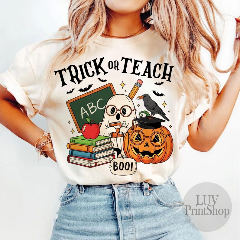 May include: Cream-colored t-shirt with a Halloween-themed graphic. The design features a ghost, a jack-o'-lantern wearing glasses, a chalkboard with "ABC", books, and the text "TRICK OR TEACH". A crow sits atop the pumpkin.