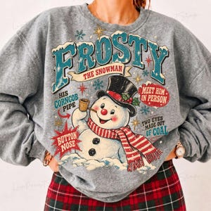 May include: A heather gray crewneck sweatshirt featuring a graphic of a snowman wearing a black top hat and a red and white striped scarf. The text reads FROSTY THE SNOWMAN, HIS CORNCOB PIPE, BUTTON NOSE, MEET HIM IN PERSON, and TWO EYES MADE OUT OF COAL.