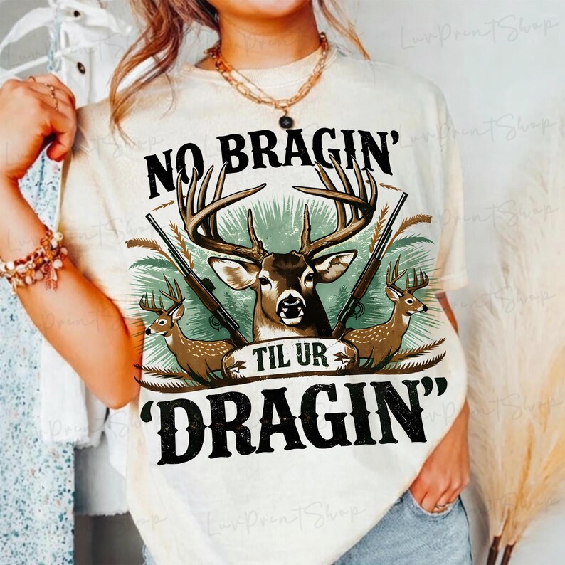 May include: Cream-coloured t-shirt with a deer graphic and the text "NO BRAGIN' TIL UR DRAGIN'". The design includes two rifles and two deer, with a central deer head. The shirt has a relaxed fit.