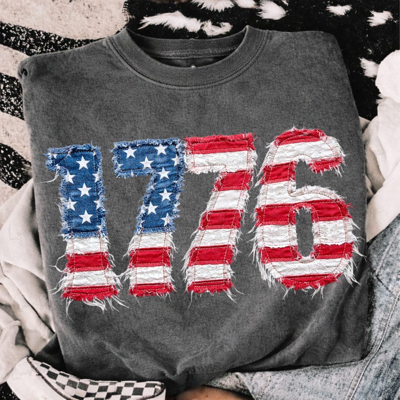 May include: A dark gray sweatshirt with the number "1776" prominently displayed on the front. The numbers are designed to resemble the American flag, with blue denim fabric featuring white stars for the "1" and "7", and red and white stripes for the "7" and "6".