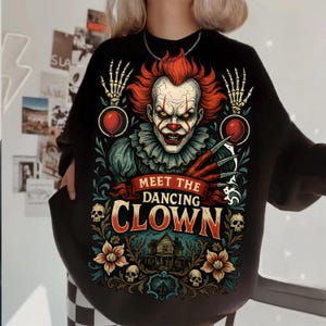May include: A black long-sleeve sweatshirt featuring a graphic print of a clown with red hair, sharp claws, and skeleton hands holding red balloons. The text reads MEET THE DANCING CLOWN above a house illustration, skulls, and floral motifs.