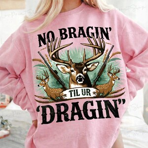 May include: Pale pink sweatshirt featuring a deer graphic and the text "NO BRAGIN' TIL UR DRAGIN'". The graphic includes a deer with antlers, rifles, and smaller deer. The sweatshirt is a light pink colour.
