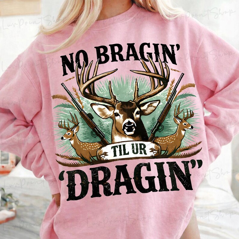May include: Pale pink sweatshirt featuring a deer graphic and the text "NO BRAGIN' TIL UR DRAGIN'". The graphic includes a deer with antlers, rifles, and smaller deer. The sweatshirt is a light pink colour.
