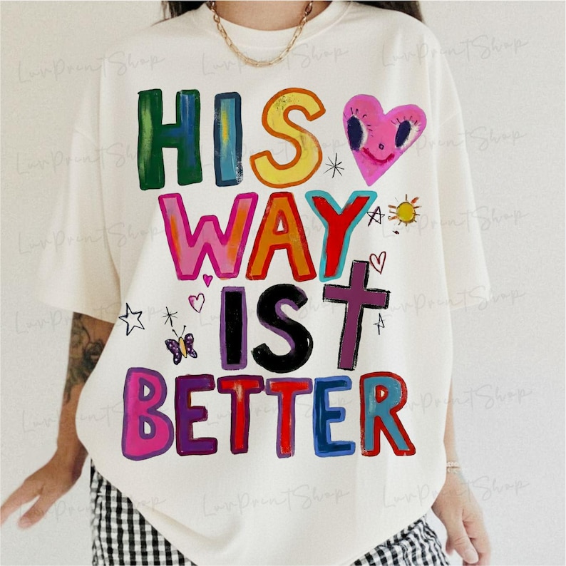 May include: Cream-colored t-shirt with the phrase "HIS WAY IS BETTER" in colorful, block letters. The design includes a pink heart, a cross, a sun, and other small illustrations. The shirt is a casual, oversized fit.
