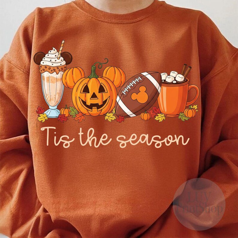 May include: Orange sweatshirt featuring a seasonal graphic. The design includes a milkshake, a Mickey Mouse-shaped pumpkin, a football, and a mug of hot chocolate. The text "Tis the season" is written in a cursive font.