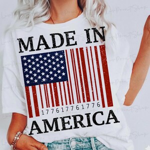 Peut inclure: T-shirt blanc avec un motif vieilli du drapeau am&eacute;ricain en forme de code-barres. Les mots "MADE IN AMERICA" sont imprim&eacute;s en noir, avec l'ann&eacute;e "1776" int&eacute;gr&eacute;e dans le code-barres.