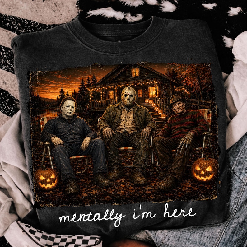 Peut inclure: Un sweat-shirt gris fonc&eacute; &agrave; col rond avec une impression graphique repr&eacute;sentant trois personnages de films d'horreur assis sur des chaises devant une cabane la nuit. Deux citrouilles d'Halloween sont au sol. Le texte "mentally i'm here" est imprim&eacute; en blanc.