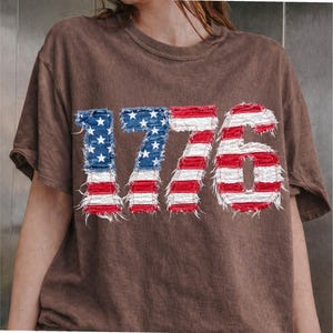 May include: Brown t-shirt featuring the number "1776" in a distressed American flag design. The numbers are in blue with white stars and red and white stripes, giving a vintage look. The shirt has a crew neck and short sleeves.