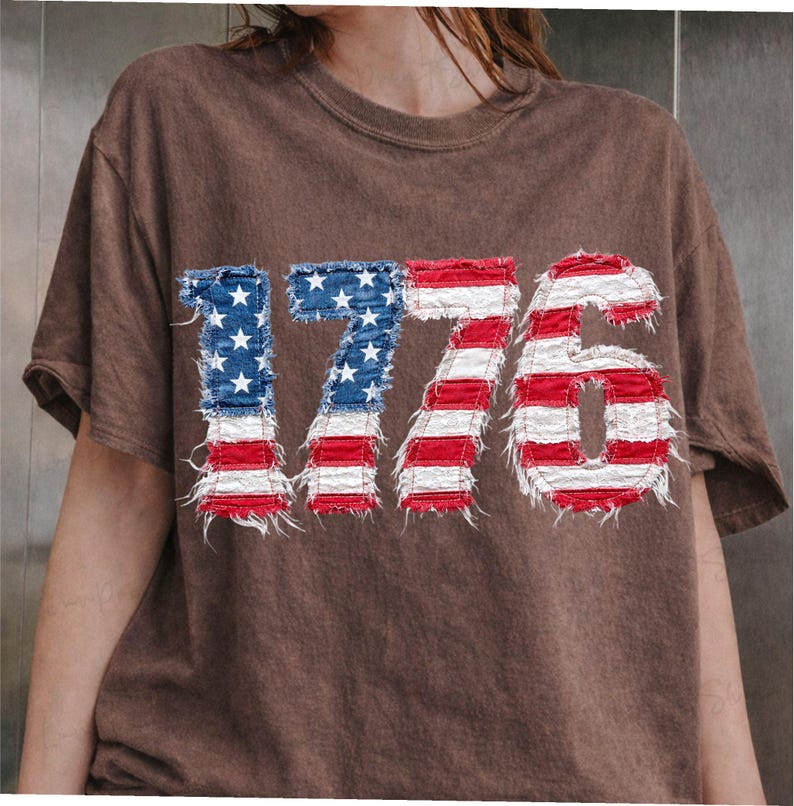May include: Brown t-shirt featuring the number "1776" in a distressed American flag design. The numbers are in blue with white stars and red and white stripes, giving a vintage look. The shirt has a crew neck and short sleeves.