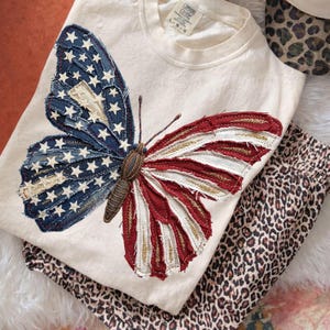 Puede incluir: Camiseta de manga larga color crema con un gran dise&ntilde;o de mariposa. Las alas de la mariposa est&aacute;n hechas de tela vaquera y tela roja y blanca, que se asemejan a la bandera estadounidense. Debajo hay un pantal&oacute;n con estampado de leopardo doblado.