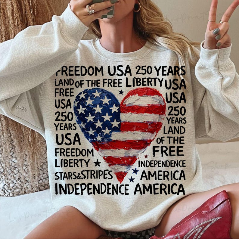 Puede incluir: Una sudadera gris claro con un dise&ntilde;o de bandera estadounidense en forma de coraz&oacute;n. La bandera est&aacute; dividida en dos mitades, una con estrellas azules y la otra con rayas rojas y blancas. El texto incluye "Freedom", "Liberty" e "Independence".