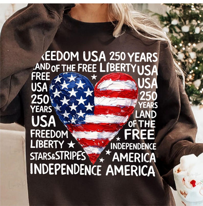Puede incluir: Una sudadera marr&oacute;n con un dise&ntilde;o en forma de coraz&oacute;n con los colores de la bandera estadounidense. El dise&ntilde;o est&aacute; dividido, con estrellas azules en un lado y rayas rojas y blancas en el otro. El texto incluye "Freedom USA 250 Years".