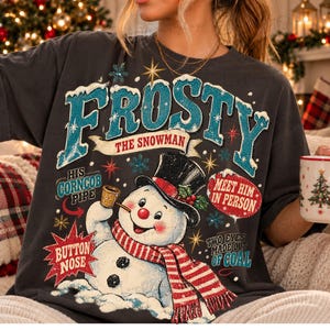 May include: A charcoal gray t-shirt featuring a graphic of a snowman wearing a black top hat and a red and white striped scarf. The text reads FROSTY THE SNOWMAN, HIS CORNCOB PIPE, BUTTON NOSE, MEET HIM IN PERSON, and TWO EYES MADE OUT OF COAL.