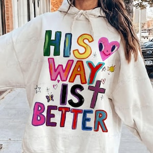 May include: Cream-colored hoodie with the text "HIS WAY IS BETTER" in colorful, block letters. The design includes a pink heart, a cross, a sun, and other small illustrations. The hoodie has a drawstring hood.